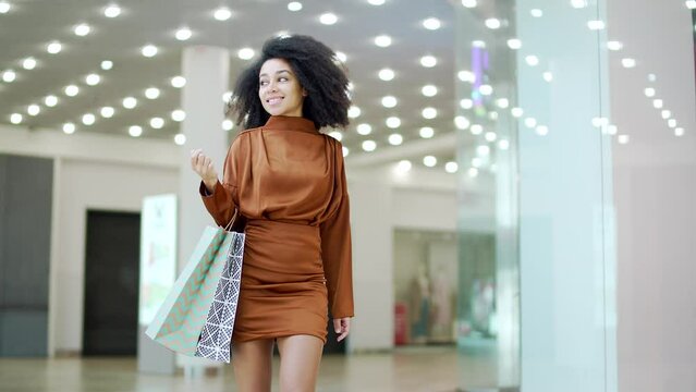 Happy young beautiful woman walking in shopping center mall between shops store. Delighted female lady in dress with shopping or gift packages bag on sale or discount day or black friday indoor plaza