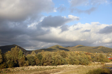 Fototapeta premium Countryside in mountains at sunrise. Grassy rural slopes with fields and trees in fall foliage in autumn. Magnificent mountain in the hazy distance.