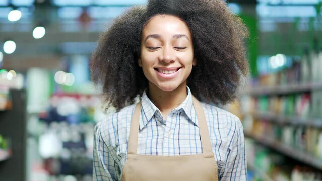 Portrait of a saleswoman female worker in a supermarket looking at camera, smiling. African american black woman employee clerk in apron in grocery store Happy Positive girl in market indoor Closeup