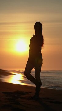 Sensuality woman in short dress, high boots dancing on sandy beach at sunrise on background solar disk rising above horizon during summer beach holiday at golden hour. Slow motion, handheld, vertical