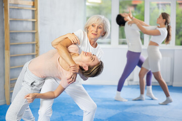 Emotional active senior woman applying inverted headlock, painful control grip to hold female opponent during mock bout in self-defense training..