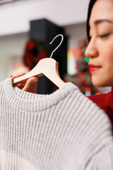 Client looking at clothing on hangers before buying presents for christmas eve winter festivity, person checking sweater material. Asian woman customer checking items on sale in store. Close up.