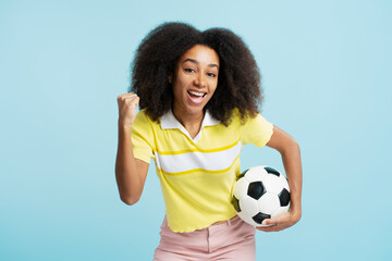 Smiling African American woman holding soccer ball, looking at camera isolated on blue background