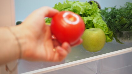 Woman hand taking, grabbing or picks up fresh fruits, vegetables and herbs out of open refrigerator shelf or fridge drawer full of fruits, vegetables, apple, tomatoes, cucumbers, lettuce. Healthy food