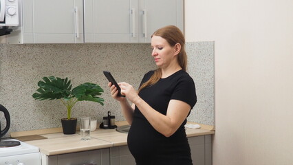 Beautiful mature pregnant woman in the kitchen home watching funny video on smartphone. Happy pregnant woman using mobile while touching tummy at home. Middle aged expecting woman using cellphone.