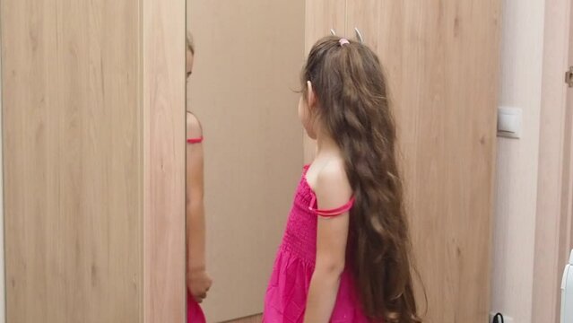 a little pretty girl with long dark hair tries on a summer pink sundress at home in front of a mirror on the closet. to go to a holiday or birthday. child fashion looking in the mirror at home