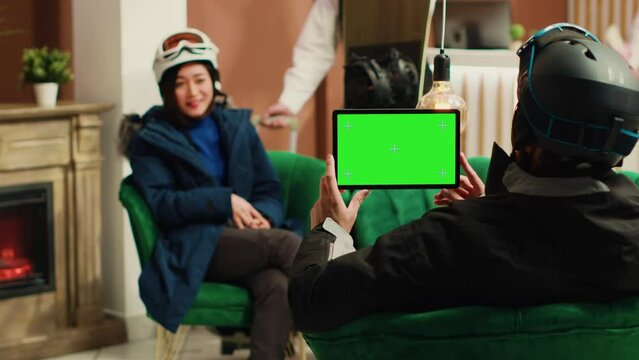 Young man uses tablet with greenscreen display in ski resort lounge area, holding digital gadget displaying blank chroma key mockup template. Couple talking about extreme winter sports.