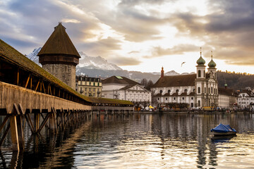 Old town of Lucerne, Switzerland at sunset in winter. Famous wooden Chapel Bridge on Reuss river, Jesuit Church, snowcapped mountain. Swiss historic buildings and medieval architecture near water - obrazy, fototapety, plakaty
