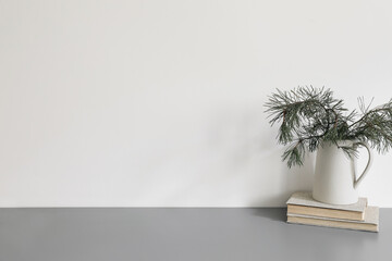 Elegant Christmas interior, home office. Ceramic vase, jug with green pine tree branches on old books. Winter home decor. Empty white wall background mockup. Grey table, desk. No people.