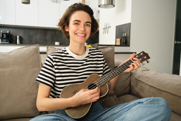 Beautiful girl playing ukulele on sofa, singing her favourite song and picking chords on small...