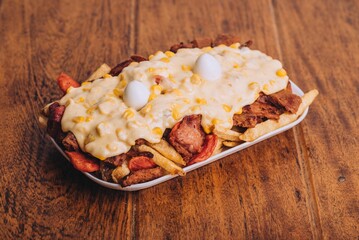 Closeup shot of a dish with french fries, sausages topped with cheese and corn on a wooden table.