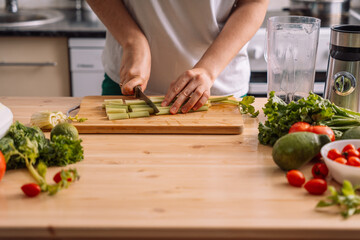 Lady in the kitchen chops celery on a wooden table on which there are fresh vegetables.