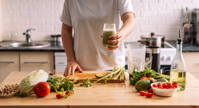 Lady in the kitchen holding a glass of smoothie in front of a wooden table on which there are fresh vegetables.