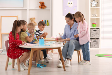 Nursery teacher and group of cute little children making toys from color paper at desks in kindergarten. Playtime activities - obrazy, fototapety, plakaty
