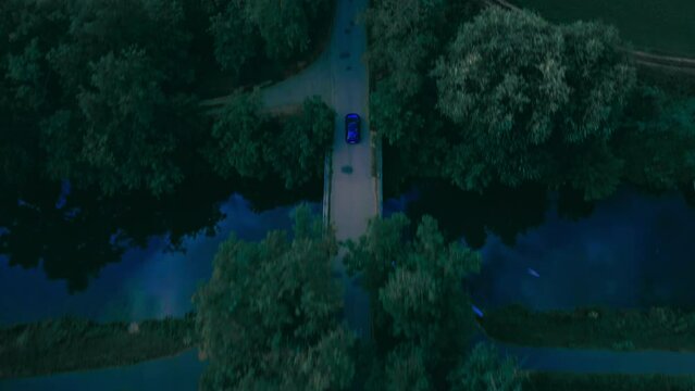 Drone footage over a blue car driving on the road with vegetation and green lawn fields