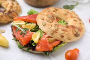 Tasty bagel with salmon, lettuce, tomatoes and arugula on white tiled table, closeup