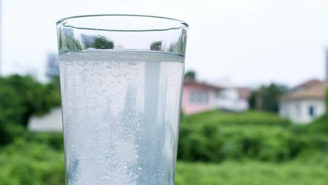Bubbly water in a glass caused by dilution of an antacid or sodium bicarbonate on a backdrop of houses and green vegetation out of focus.