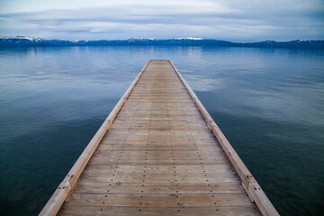 Wooden dock over the calm blue waters of Lake Tahoe with snow capped mountains in the distance