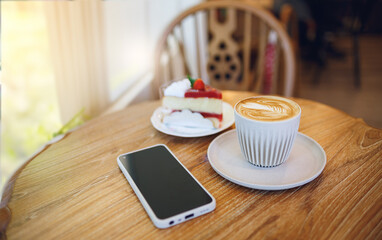 Latte coffee in cup and a piece of cherry cake with mobile phone on wooden table  by bright window and sunlight in the morning