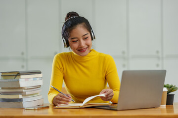 Happy Asian female student wearing wireless headphones studying online, listening to music, dancing, browsing websites, searching for information, chatting online on laptop in business study office.