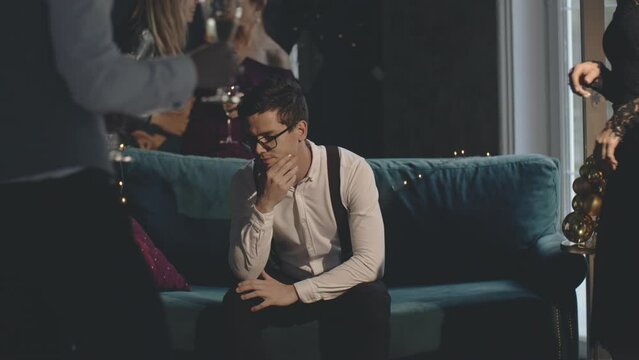 Portrait of bored and sad young man sitting alone at noisy night party. Lonely upset guy hangover with broken heart at celebration in luxury glamorous club. Single man social life. Night life concept