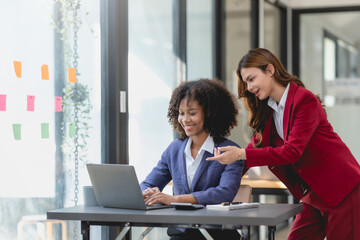 Two happy businesswoman talking and consulting working together in the office. Businesswoman discussing with colleague over laptop at desk in office.