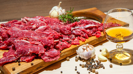 A wooden cutting board with red cube meat decorated with garlic and spices next to a glass of wine