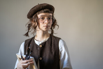 Serious illustrator in vintage clothes holding canvas, paint brushes, looking at camera. Young...