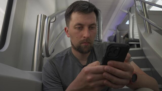 Young man travels on double decker train and sits on flyer using his smartphone. No available seating during railroad trip. Male commutes in dual deck train seated on flyer and surfing internet. 