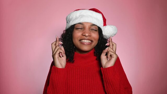 Portrait of pleading young Santa dark skinned woman in red sweater and Christmas hat making wish, posing isolated over pink color background studio