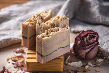 Delicate cute pieces of natural soap on a wooden soap dish on a background of fabric and pink flowers and petals nearby. Beautiful postcard