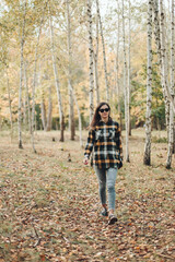 Fototapeta premium A beautiful girl in jeans and a plaid shirt walks in a birch grove. Portraits of a brunette girl in nature, in the autumn forest