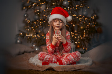A portrait of a sweet beautiful smiling little 7-old girl wearing red pyjamas and Santas hat holding glass snow globe in her hands at home near Christmas tree at Christmas eve. The concept of New Year