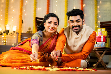 Happy indian couple in traditional wear looking camera while decorating rangoli with flower for festival celebration on floor - concept of religious custom, bonding, and religious ceremony.