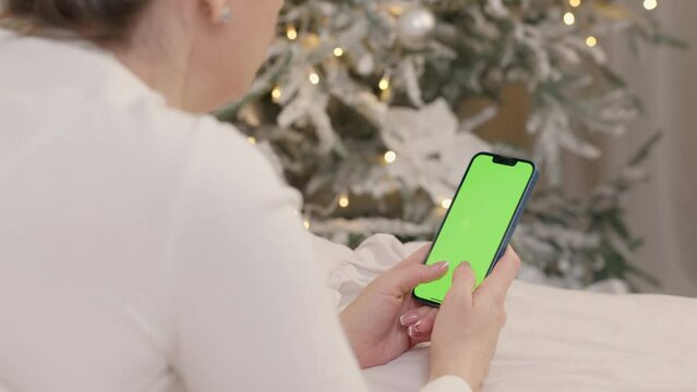 Woman Uses Phone With Green Mock-up Screen Lying On Bed In Cozy Room With Christmas Tree And Lights. Girl Surfing Internet, Browsing Social Networks and Making Online Shopping