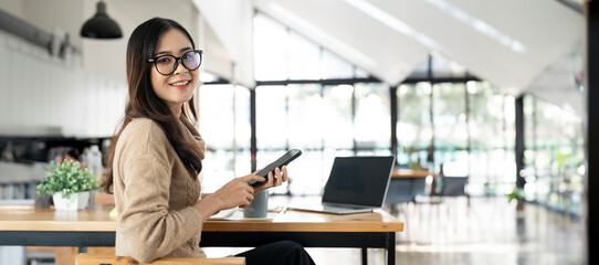 Smiling businesswoman using her phone in the office. Small business entrepreneur sitting at office and looking at camera - obrazy, fototapety, plakaty