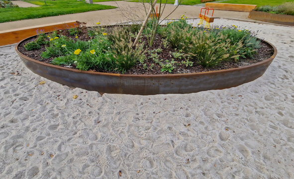 the oval ellipse of the flower bed is lined with a rusty sheet metal border. perennials are blooming. sandpit with park features and children's playground. rounded edge, stepping stones, concrete