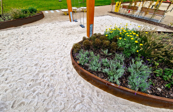 the oval ellipse of the flower bed is lined with a rusty sheet metal border. perennials are blooming. sandpit with park features and children's playground. rounded edge, stepping stones, concrete