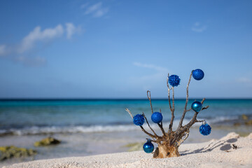 Dead corals decorated with blue Christmas balls standing on the sand beach with blue sea behind it 