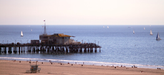 View of the Santa Monica Pier in Santa Monica, California in late afternoon. The restaurant at the end of the pier is visible, as are numerous sailboats in the Pacific Ocean and beachgoers.