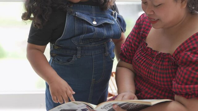 mother and girl are reading a book in living room. Asian chubby family relaxing with education program.