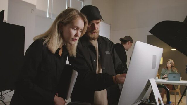 Medium shot of male production director and female manager standing together in studio, reviewing filmed footage on computer screen and discussing something, while videographer is preparing camera