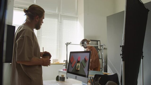 Medium shot of female photographer shooting flatlay of geometric shapes and cubes with camera on horizontal rack, and male colleague looking at result on computer screen