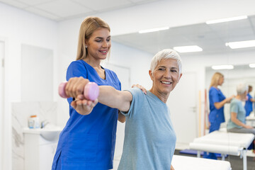 Fototapeta premium Oder aged woman doing physiotherapy with support from physiotherapists . Senior elderly female sitting in clinic using dumbbells workout exercise for patient with caregiver in nursing care.