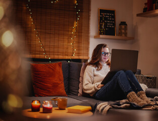 A young woman spends a cozy winter evening at home lying on the sofa with a laptop. Winter holidays, Christmas and online surfing concept