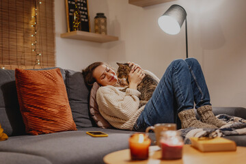 Cozy at home with tabby cat, woman with her pet on sofa ay home in evening - obrazy, fototapety, plakaty