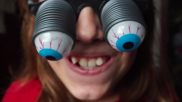 A 9-year-old caucasian girl wearing glasses with eyeballs falling out on springs. A child is preparing for Halloween and trying on funny, scary accessories. Masquerade decoration. Black humor