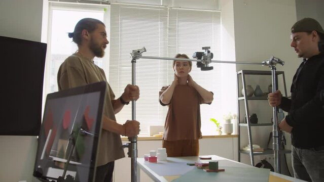 Medium shot of two male colleagues helping female photographer raise up camera mounted on telescopic rack, while preparing to shoot flatlay composition with geometric shapes set up on tableMedium shot
