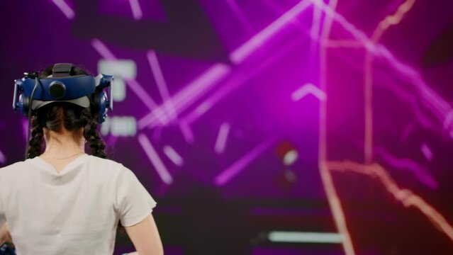 The little girl wearing VR headset plays game. The child use tracking controllers in her hands, moves actively immersing herself in modern game. Behind girl there is screen with blur background image