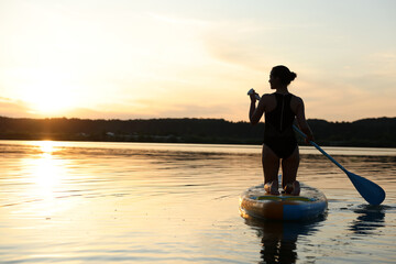 Woman paddle boarding on SUP board in river at sunset, back view